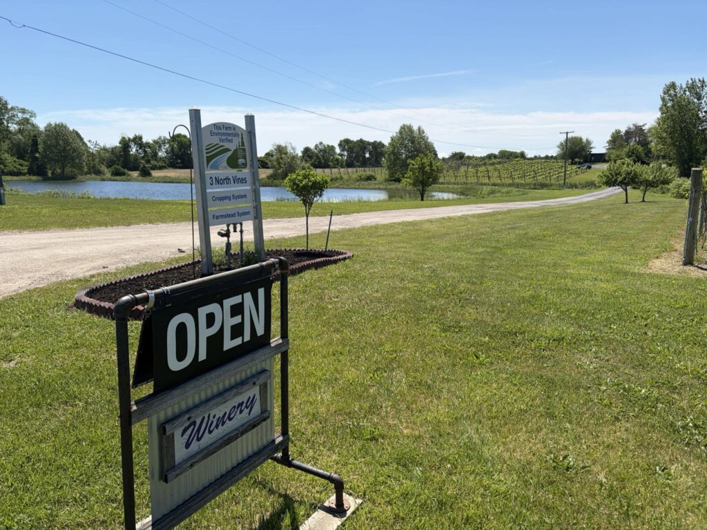 Winery entrance with "Open" sign, grassy area, pond, and vineyard in the background.