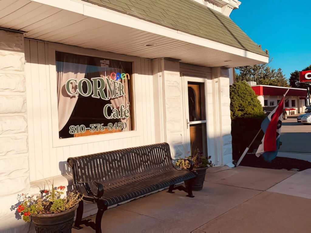 Exterior of Corner Café with a bench and flower pots outside. A sign displays the phone number, and an open sign is visible in the window. A flag is placed near the entrance.