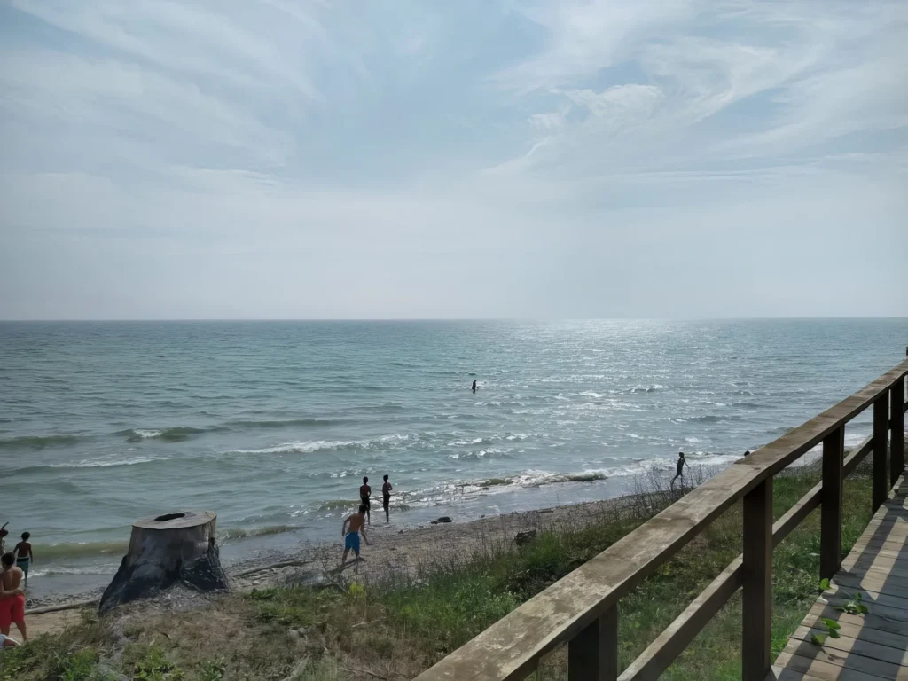 People wading in calm Lake Huron waters at a beach with wooden boardwalk railing in foreground under cloudy sky at Forester Park.