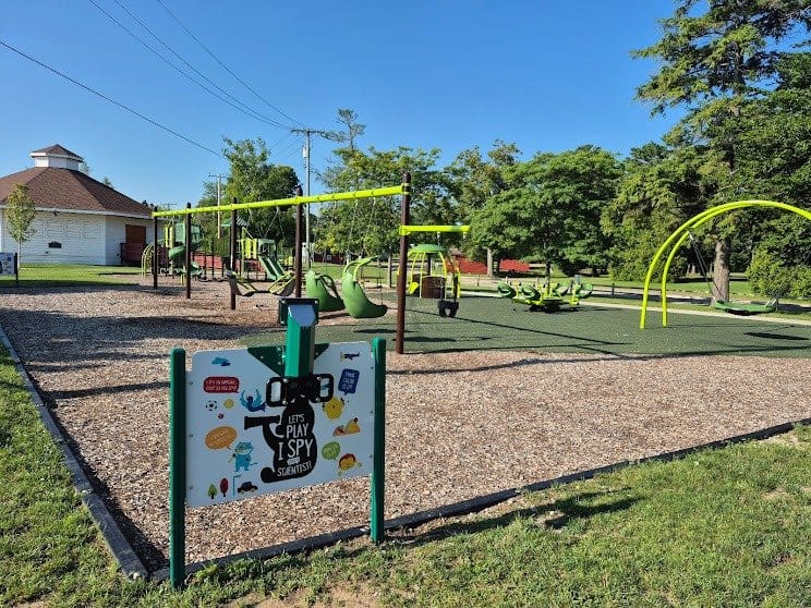 Playground at Forester Park with yellow play equipment, swings, and gravel surface surrounded by green grass and trees under blue sky.