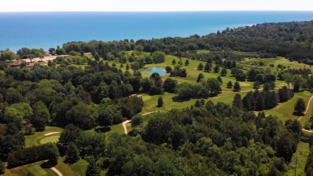 Aerial view of a lush green golf course with a small pond, surrounded by dense trees, overlooking a blue Lake Huron in the background.