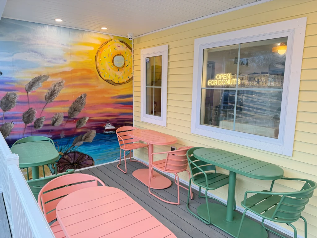 Outdoor seating area with pastel green and pink tables and chairs. A mural on the wall depicts a sunset, large donut, and plants. A window displays a neon sign reading "Open for Donuts."