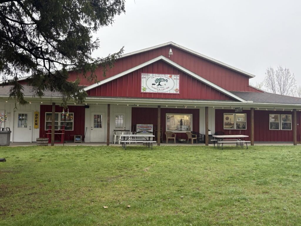 A red barn-style building with a sign for an orchard and cider mill. The front porch has tables, chairs, and an "OPEN" sign. The grass lawn is in the foreground.