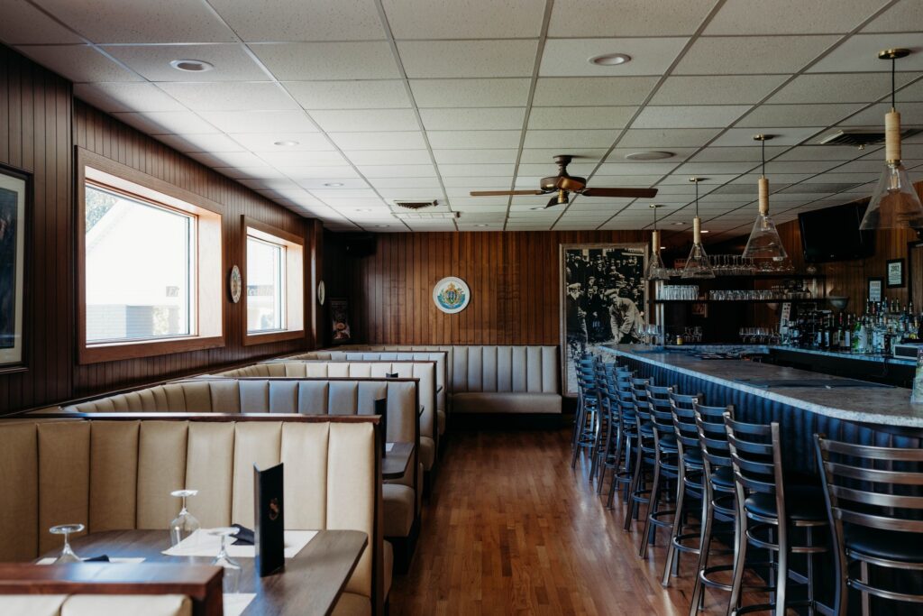 Interior of a restaurant with wooden paneling, featuring booth seating on the left and a bar with high stools on the right. Large windows provide natural light.