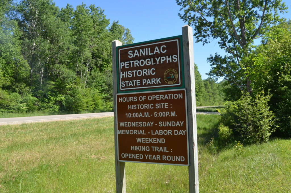 Sign for Sanilac Petroglyphs Historic State Park with hours of operation and hiking trail information, surrounded by green trees and grass.