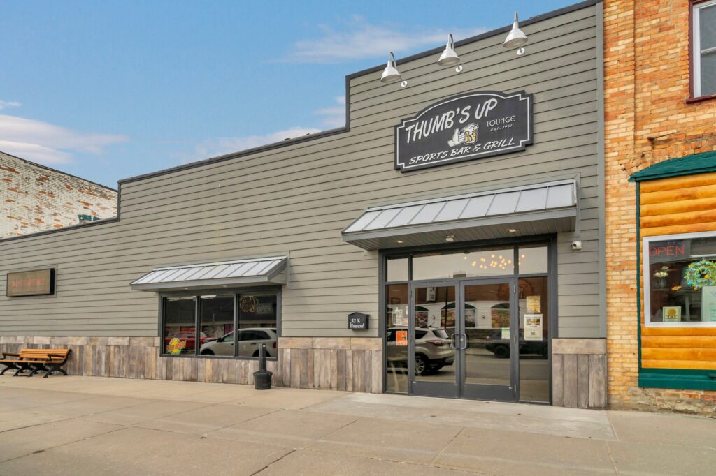 Exterior of Thumb's Up Sports Bar & Grill with gray siding, large windows, and a sign above the entrance. A bench is on the sidewalk.