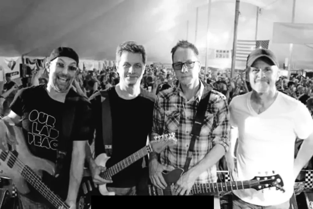 Four men holding guitars stand together on stage in front of a large crowd under a tent.