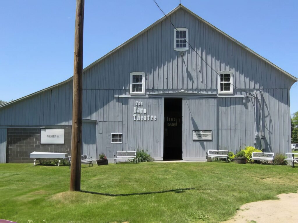 A gray barn converted into a theater, with a sign reading "The Barn Theatre" on the front. There are benches and plants outside on a grassy lawn under a clear blue sky.
