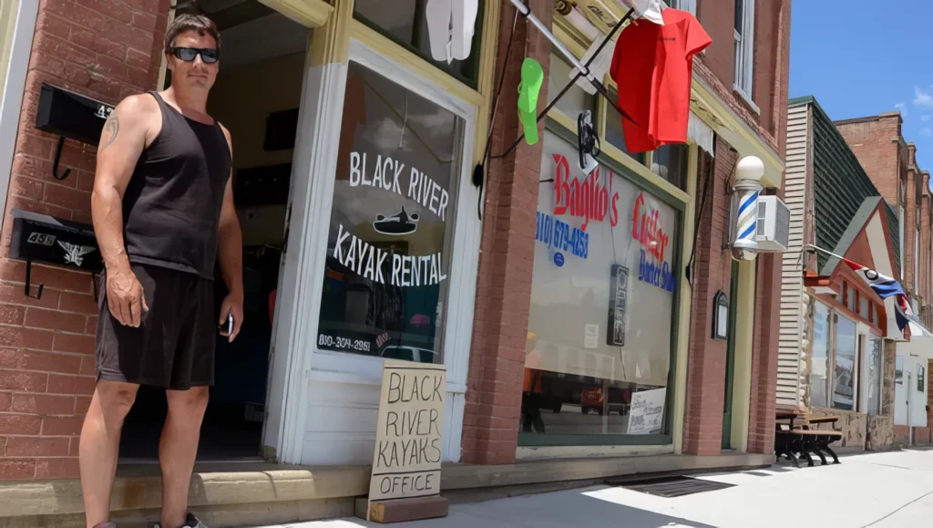 Man in sunglasses standing outside Black River Kayak Rental shop on a sunny day.