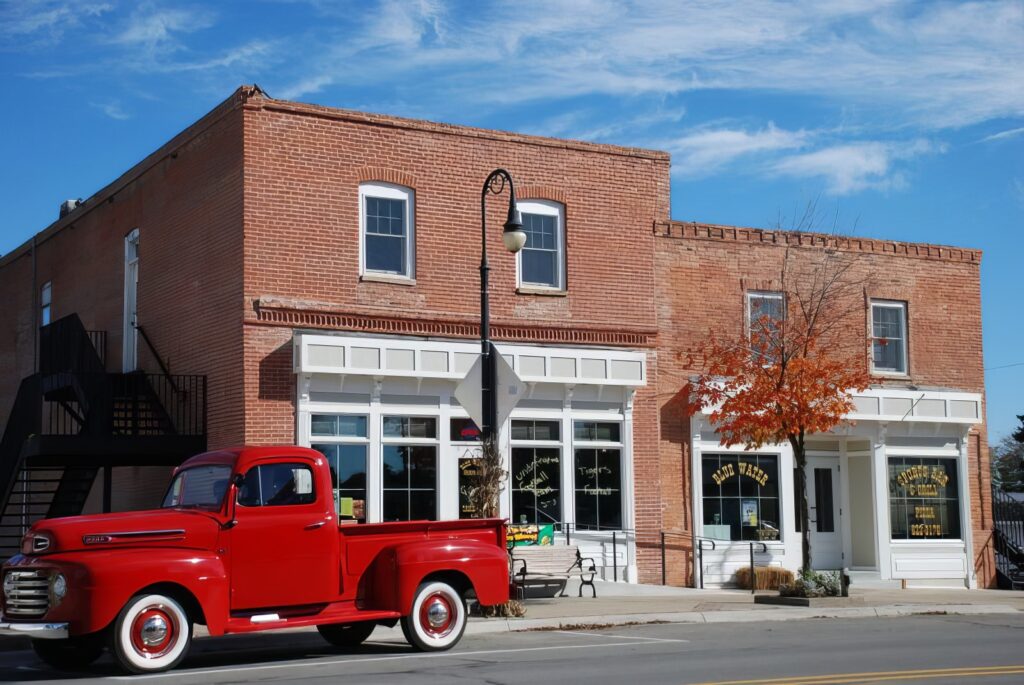 A vintage red pickup truck parked in front of two brick buildings with large windows and a small tree with orange leaves on the sidewalk.