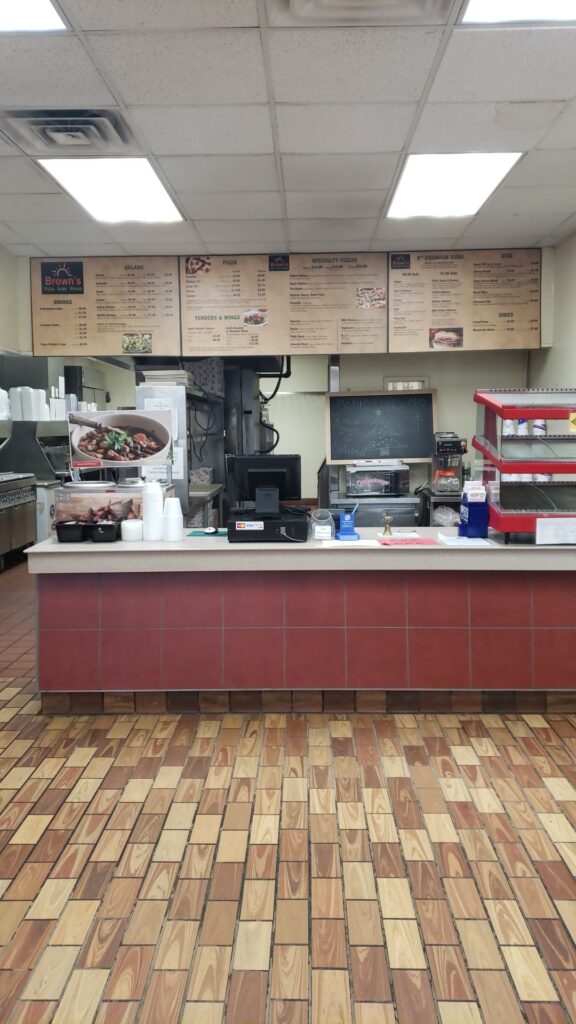 Counter of a fast-food restaurant with a menu board above, a cash register, and food displays. The floor has a wood-like tile pattern.
