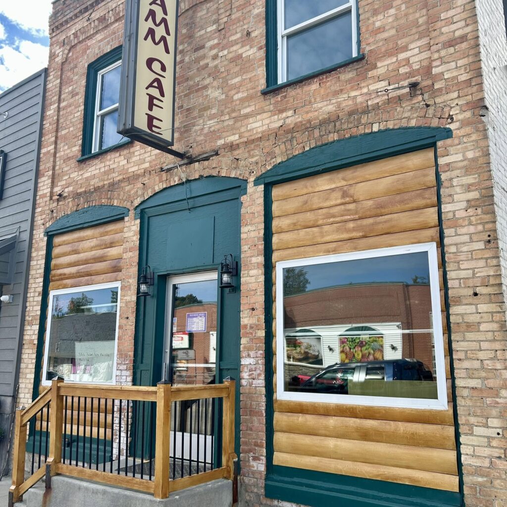 Exterior of a brick building with green trim, featuring a sign that reads "Café" and large windows reflecting a nearby building.