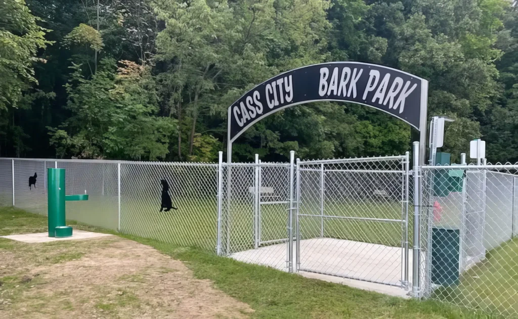 Entrance to Cass City Bark Park with a chain-link fence, surrounded by trees.