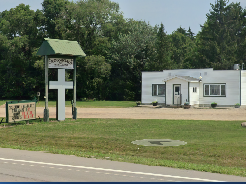 A small white building with a green roof sign labeled "Crossroads Restaurant" next to a grassy area and trees.