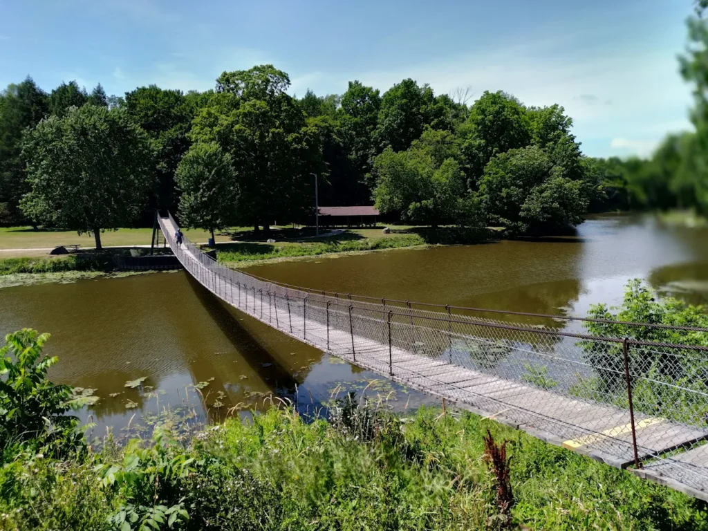 The Croswell Swinging Bridge crosses over a calm river, surrounded by lush green trees and vegetation under a clear blue sky.