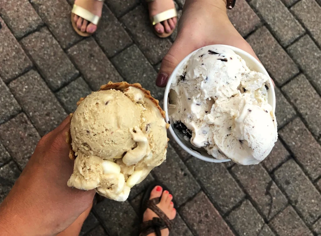 Two people holding ice cream; one in a waffle cone and the other in a cup, standing on a brick pavement.