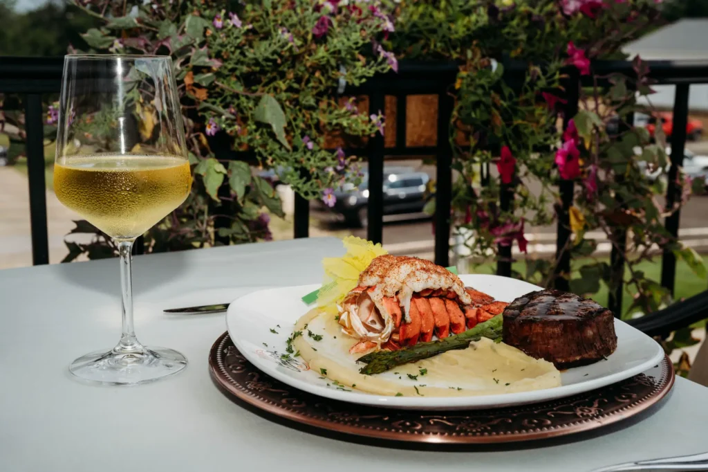 A glass of white wine next to a plate of lobster tail, steak, asparagus, and mashed potatoes on an outdoor table with plants in the background.