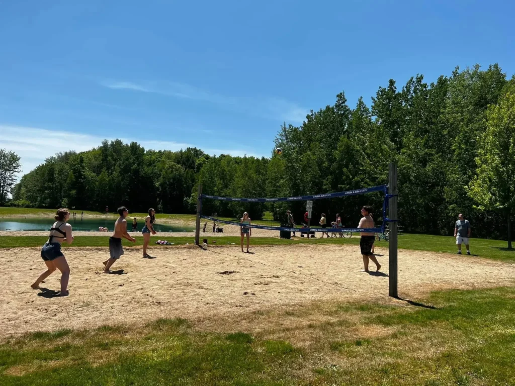 People playing beach volleyball on a sand court near the swimming pond at Evergreen Park with trees in the background.