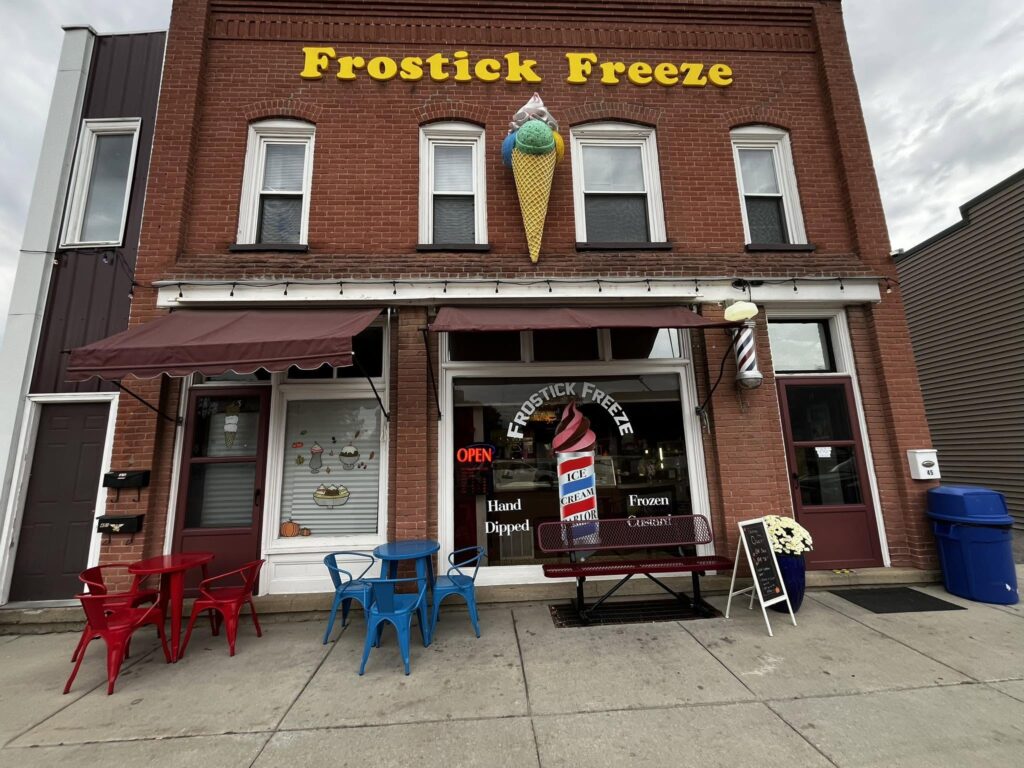 Front of Frostick Freeze, a brick ice cream shop with a large cone decoration above the door. Red and blue outdoor seating is available, with a small chalkboard sign and a trash bin nearby.