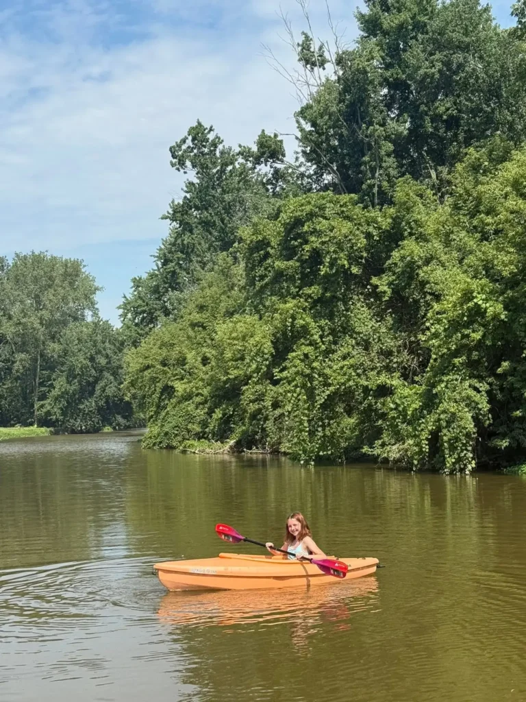 Young girl kayaking in an orange kayak with a pink paddle on the calm Cass River surrounded by lush green trees at Evergreen park
