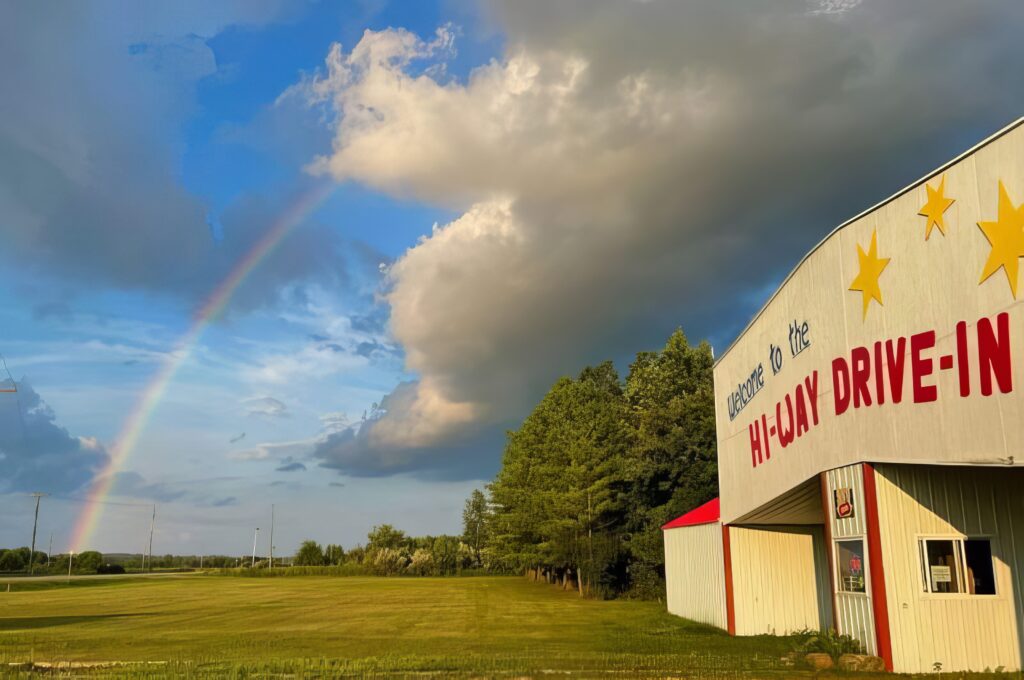 A rainbow arcs across a partly cloudy sky above a grassy field. On the right, a building with a sign reads "Welcome to the Hi-Way Drive-In," adorned with yellow stars.