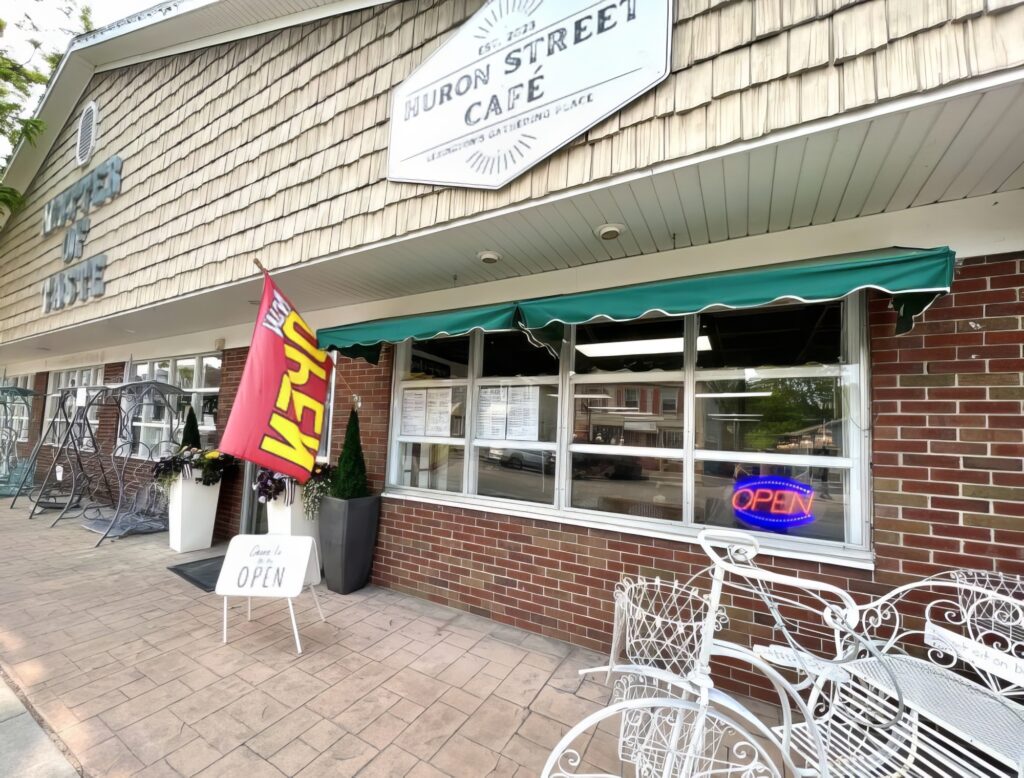 Exterior of Huron Street Cafe with a red "Open" flag, a neon "Open" sign in the window, and a white sandwich board sign on the sidewalk. White metal chairs and tables are in front.