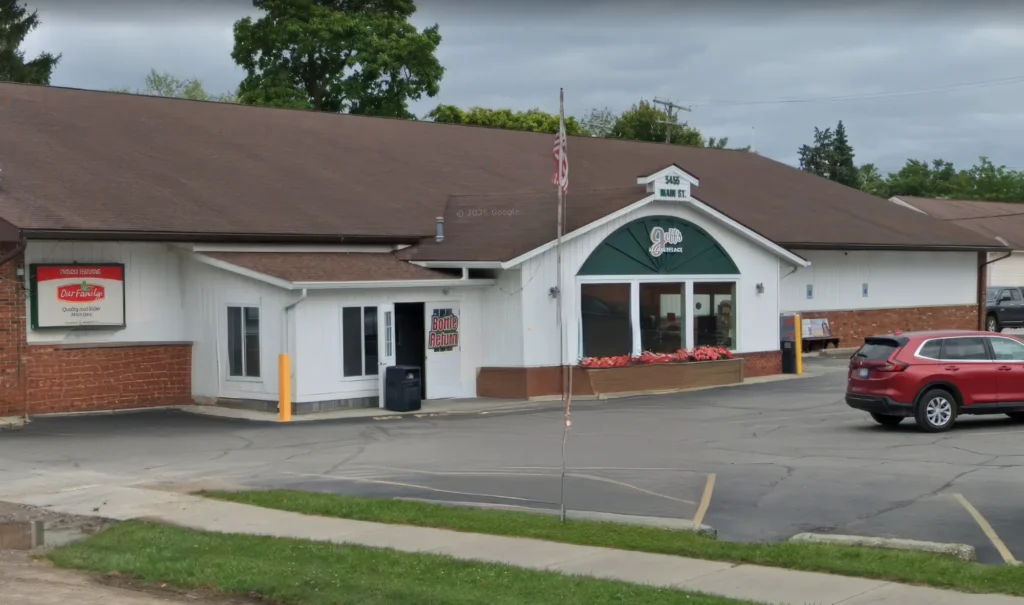 Exterior view of a grocery store with a brown roof and white siding. An American flag is displayed in front, and a red SUV is parked in the lot. Signs for "Bottle Return" and "Our Family" products are visible.