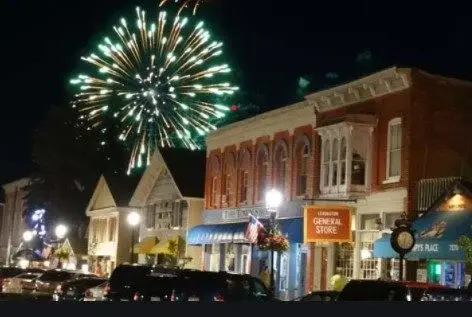 Fireworks light up the night sky over a quaint town street with lit storefronts and parked cars.