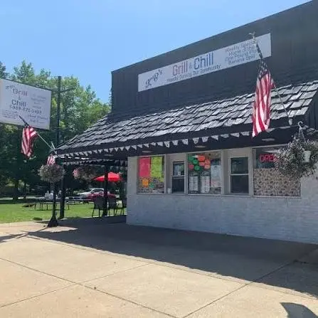 Small diner with a black roof and white walls, displaying American flags. Bright signs and an "Open" sign are visible on the front. Outdoor seating with red umbrellas is in the background.