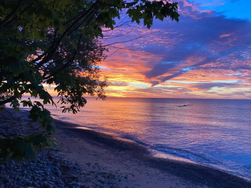 Sunset over a calm Lake Huron with vibrant orange, pink, and purple clouds, silhouetted trees on the left, and a sandy shoreline at Lexington Park.