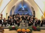 An orchestra seated in a semi-circle inside a church, with musicians holding various string instruments. The setting features arched ceilings and floral decorations in the foreground.