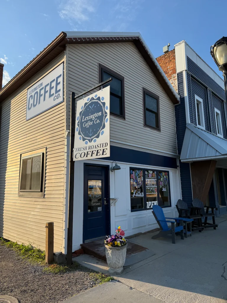 Exterior of Lexington Coffee Co., a small coffee shop with a beige and navy facade, a sign reading "Fresh Roasted Coffee," and outdoor seating with blue chairs.