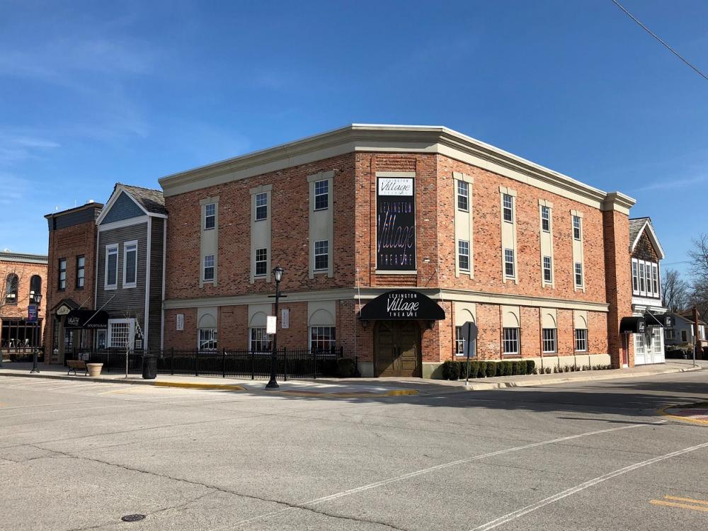 A large brick building with a sign reading "Village Theatre" on the corner, featuring multiple windows and a clear blue sky background.