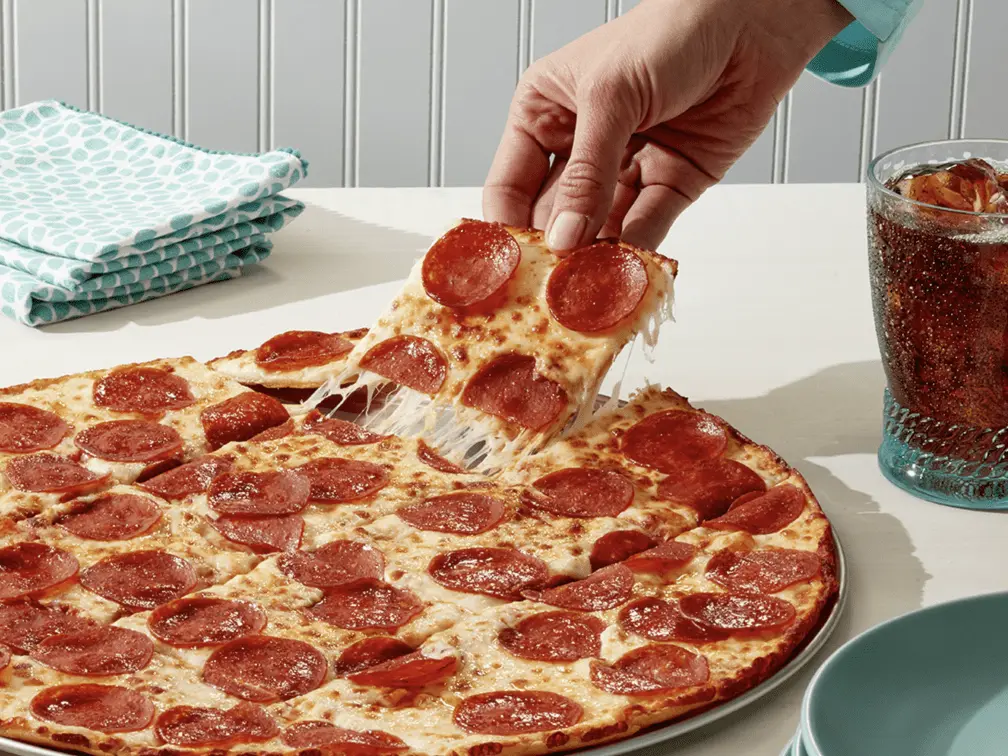 A hand lifting a slice of pepperoni pizza with melted cheese from a whole pizza on a table. A glass of soda and folded napkins are nearby.