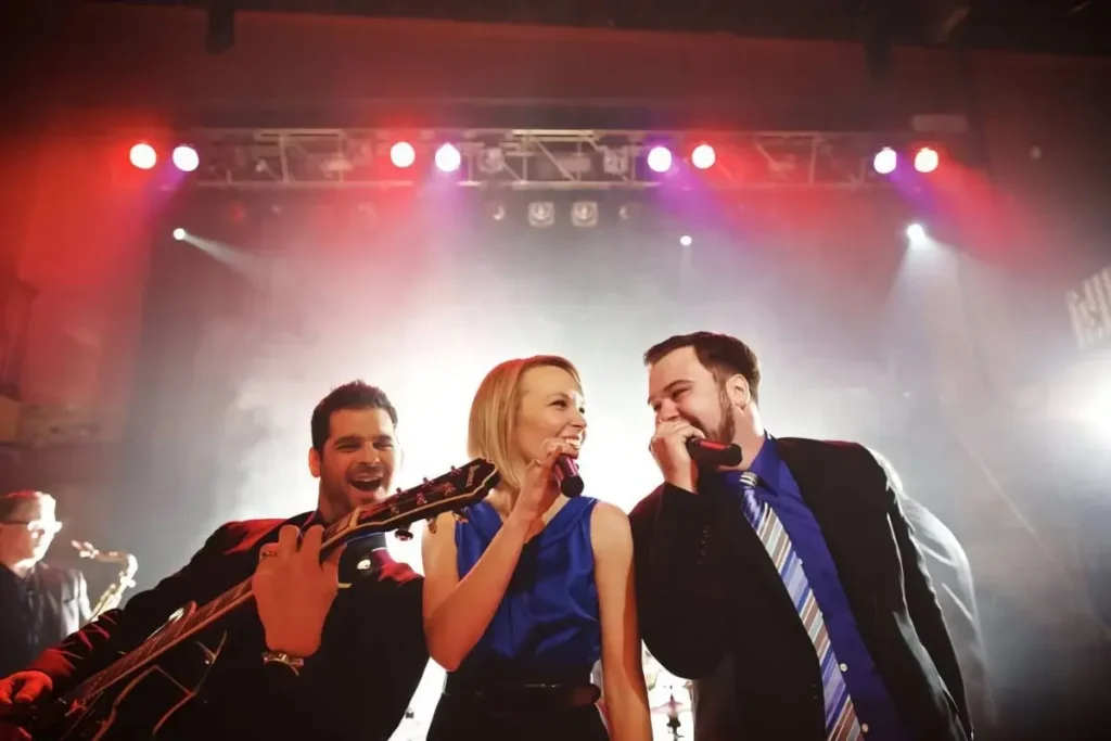 A band performing on stage with a guitarist and two singers under colorful stage lights.