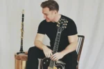 A person in black clothing sits on a chair holding a Gretsch guitar, with a clarinet resting on a nearby table.
