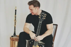 A person in black clothing sits on a chair holding a Gretsch guitar, with a clarinet resting on a nearby table.