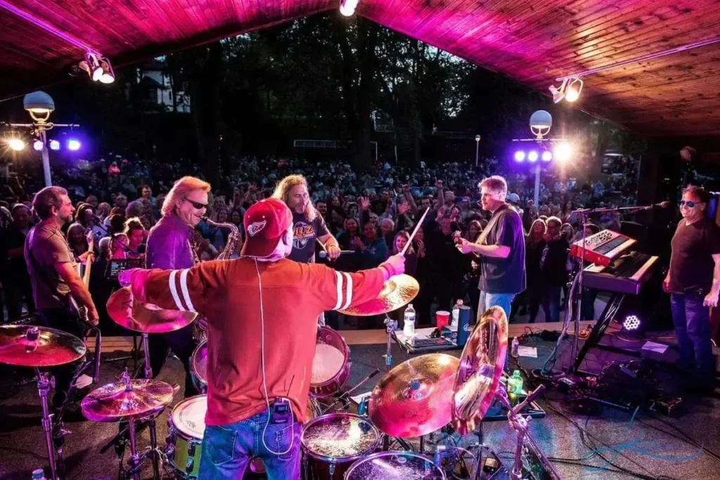 A band performs on an outdoor stage with a drummer in the foreground, surrounded by a large, cheering crowd under colorful stage lights.