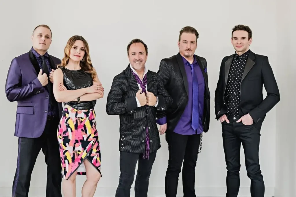 A group of five people dressed in formal and colorful attire, standing against a plain background.