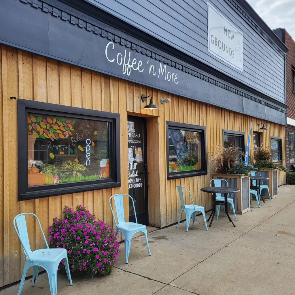 Exterior of a café named "Coffee 'n More" with wooden siding, decorated windows, and blue metal chairs outside. A sign reads "Happy Fall, Open."