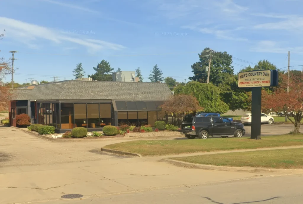 Exterior view of Nick's Country Oven, a family restaurant with a sign advertising breakfast, lunch, and dinner. A black truck is parked nearby, surrounded by landscaped bushes and trees.