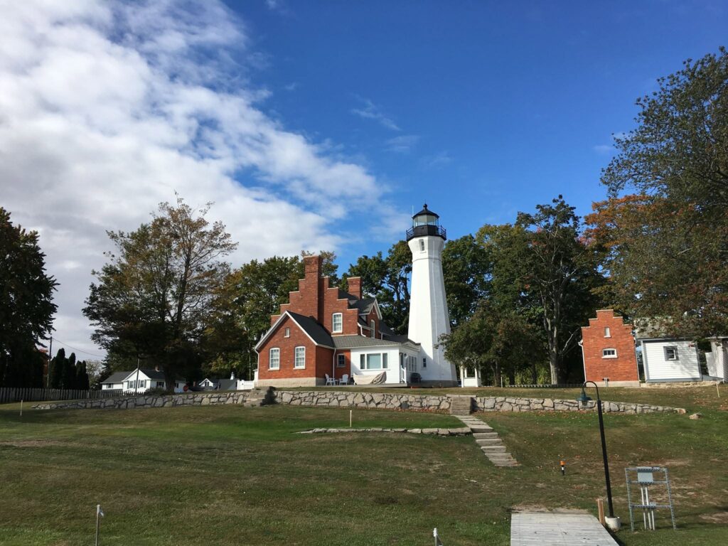A red brick house and a white lighthouse surrounded by trees under a partly cloudy sky.