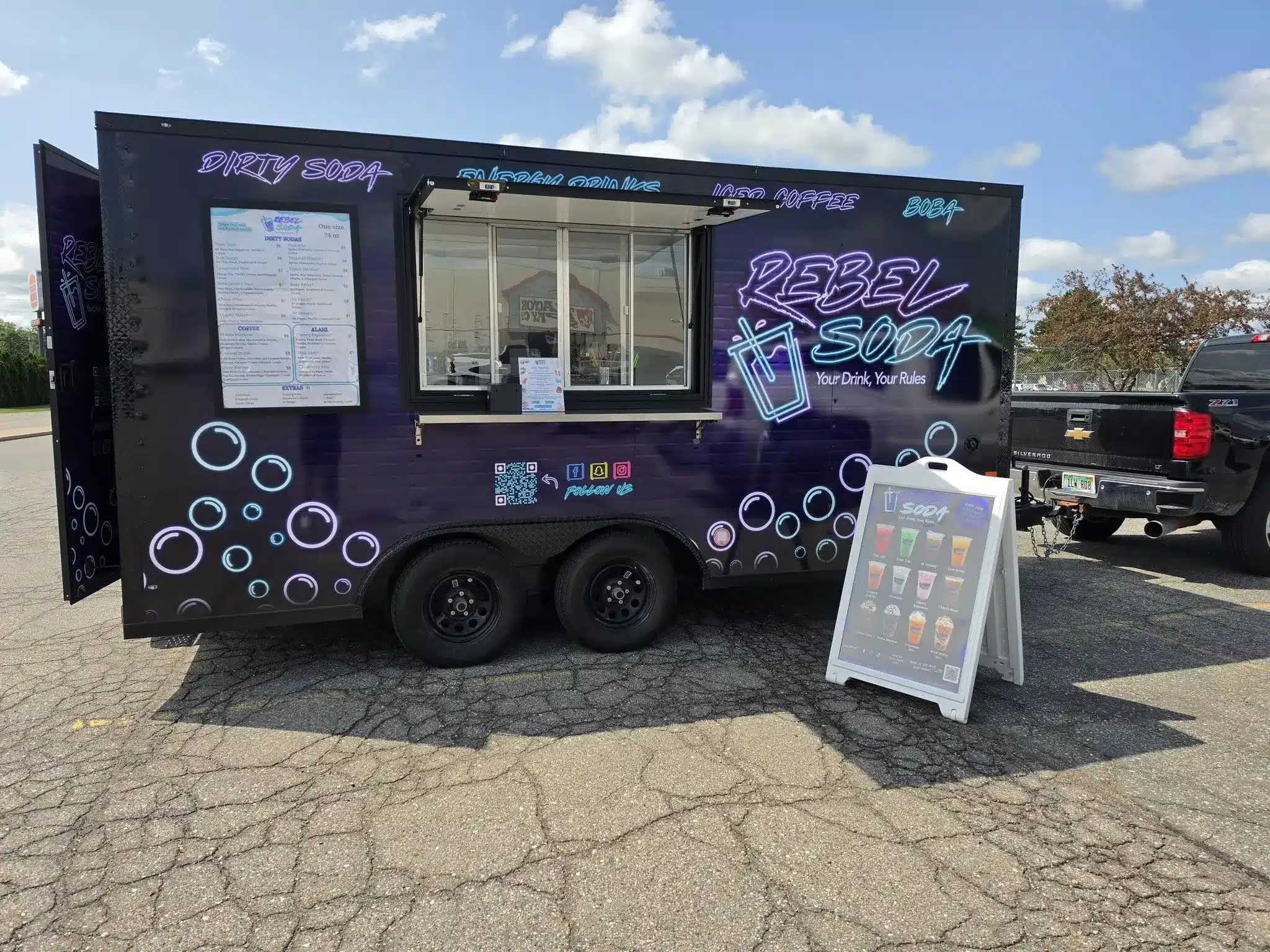 A colorful food truck named "Rebel Soda" with a menu displayed on the side. The truck features vibrant graphics and a window for serving drinks. A sign in front lists various beverage options. The setting is an open parking area with a blue sky and scattered clouds.