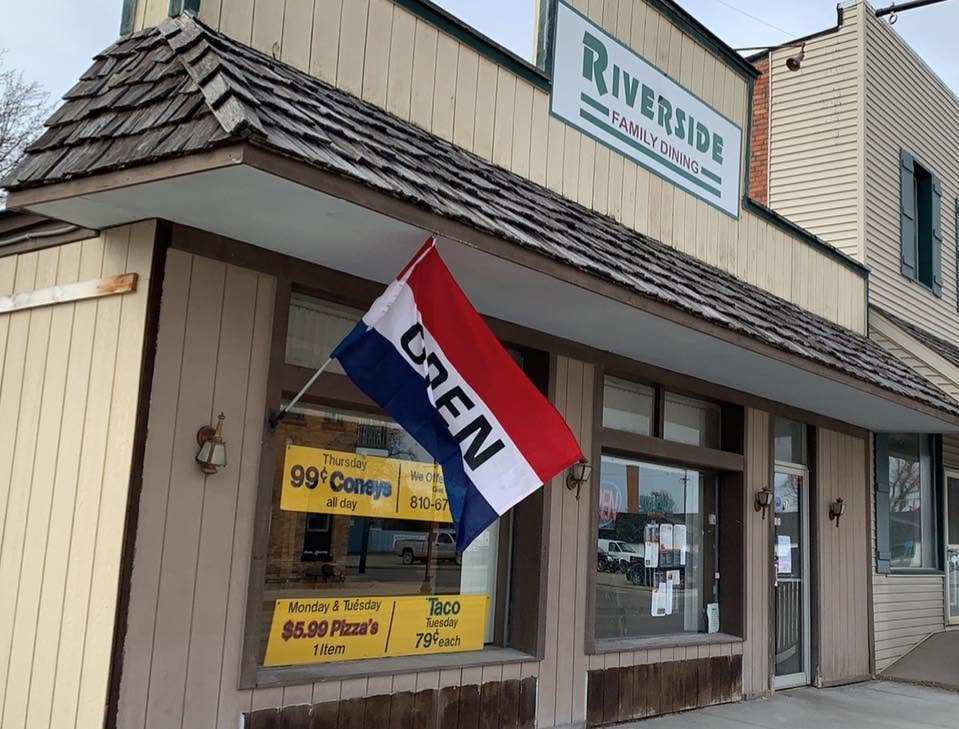 Exterior of Riverside Family Dining with an "Open" flag. Signs in the window advertise specials like 99-cent coneys and $5.99 pizzas.