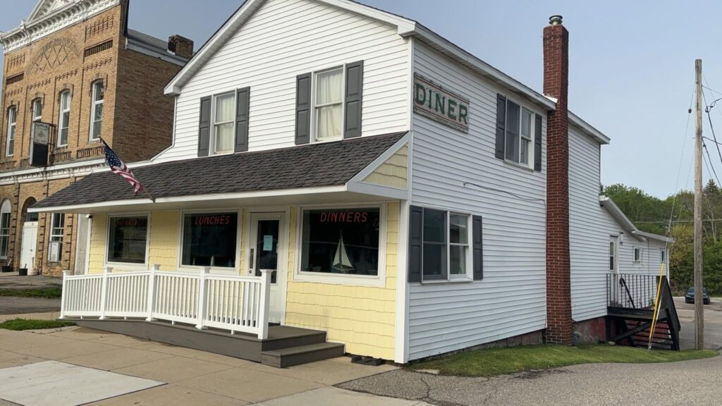 A small white and yellow diner with a front porch and an American flag. The building has signs for breakfast, lunches, and dinners.