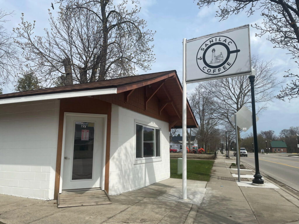 Exterior of a small white building with a sign reading "Sanilac Coffee" on a pole, located on a sidewalk with trees and a street in the background.