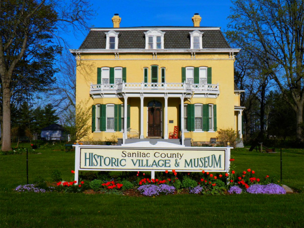 Yellow historic house with green shutters, surrounded by trees and flowers. A sign in front reads "Sanilac County Historic Village & Museum."