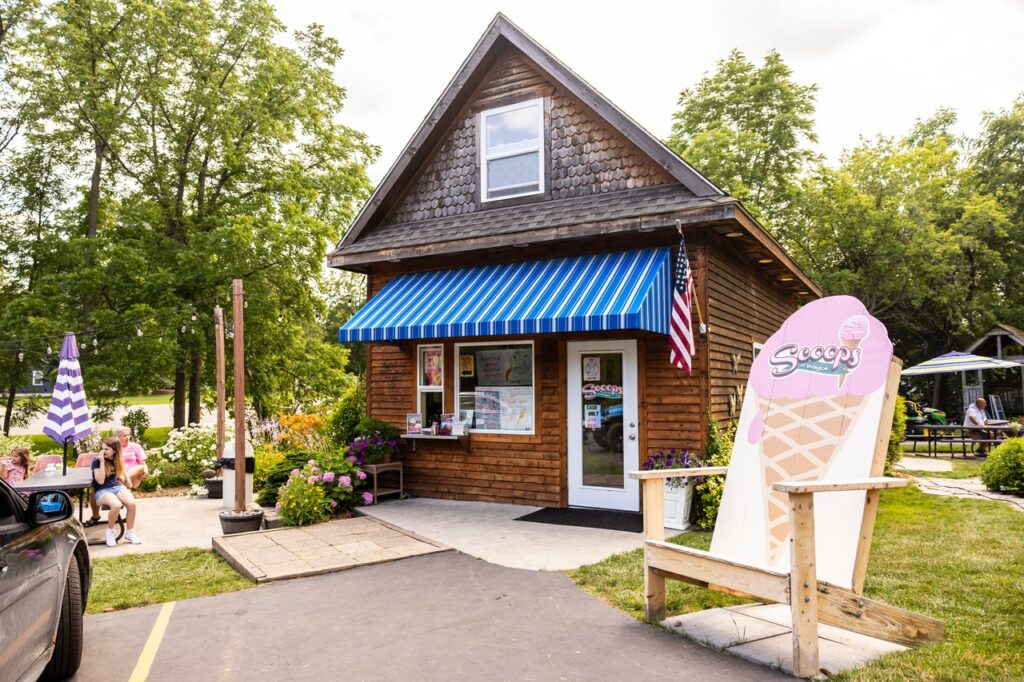 A small wooden ice cream shop with a blue striped awning and an American flag. A large ice cream cone sign is displayed outside. People are sitting at a picnic table under a purple umbrella.