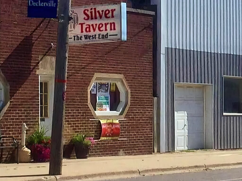 Exterior of a brick building with a sign reading "Silver Tavern - The West End." A window displays posters, and there are potted plants on the sidewalk.