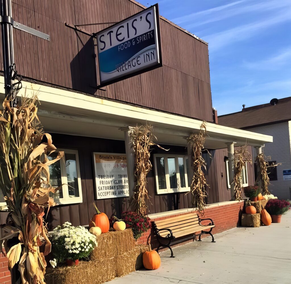Exterior of a rustic building with a sign reading "Steis's Village Inn." The entrance is decorated with cornstalks, hay bales, pumpkins, and potted flowers, creating an autumnal theme.